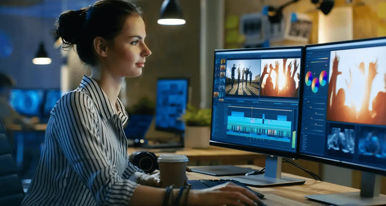 A young woman with dark hair in a bun sits at a dual-monitor editing workstation in a dimly lit creative studio, focused on a video editing timeline with colorful clips and footage of a silhouetted crowd. A coffee cup and camera strap rest nearby, representing an offshore video editor delivering polished content for a remote-first media or marketing team.