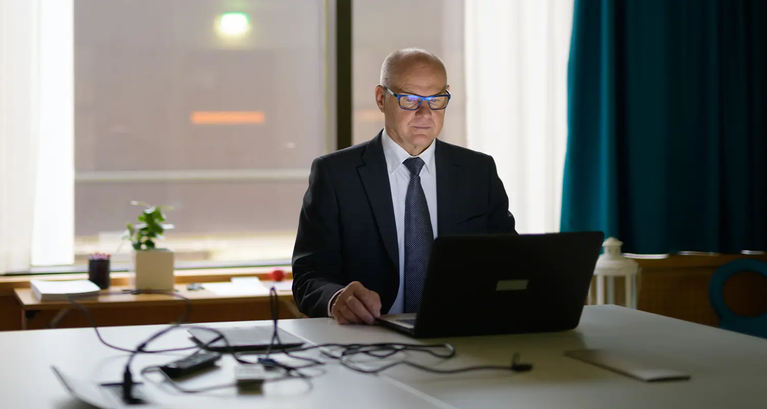 An older male executive in a dark suit and tie sits alone at a wide conference table in a dimly lit room, focused on a laptop. Blue light from the screen reflects off his glasses as he works late, illustrating the focused, experienced leadership style that global senior executive hires bring to distributed organizations.