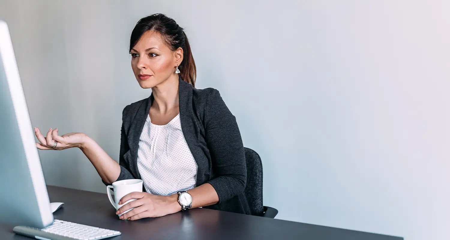 A professional woman with dark hair pulled back in a ponytail sits at a sleek black desk holding a coffee mug, looking toward a computer monitor with a calm, confident expression and an open-handed gesture. She represents a senior global executive engaging thoughtfully in a remote leadership context.