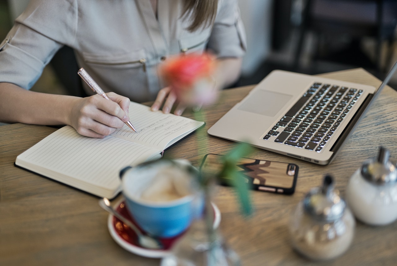 A woman's hand writing in an open lined notebook with a sparkly pen at a cafe table. A laptop, smartphone, coffee cup, and small vase with a flower are also visible on the wooden table, depicting a content writer working in a casual setting.