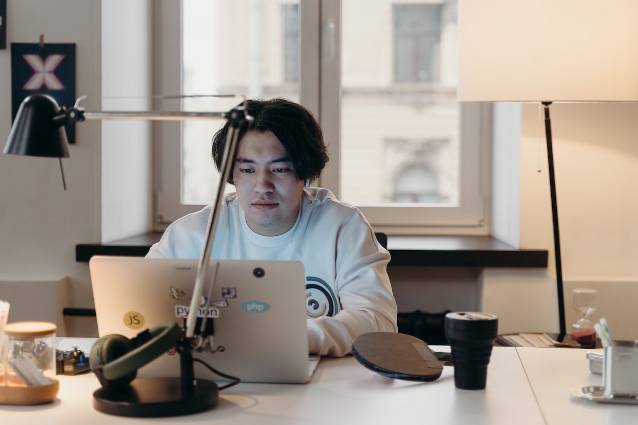 A young man with dark hair wearing a light hoodie sits focused at a desk with a laptop covered in programming stickers including JavaScript, Python, and PHP logos. A ring light, headphones, and desk lamp are visible in the dimly lit home office setup.