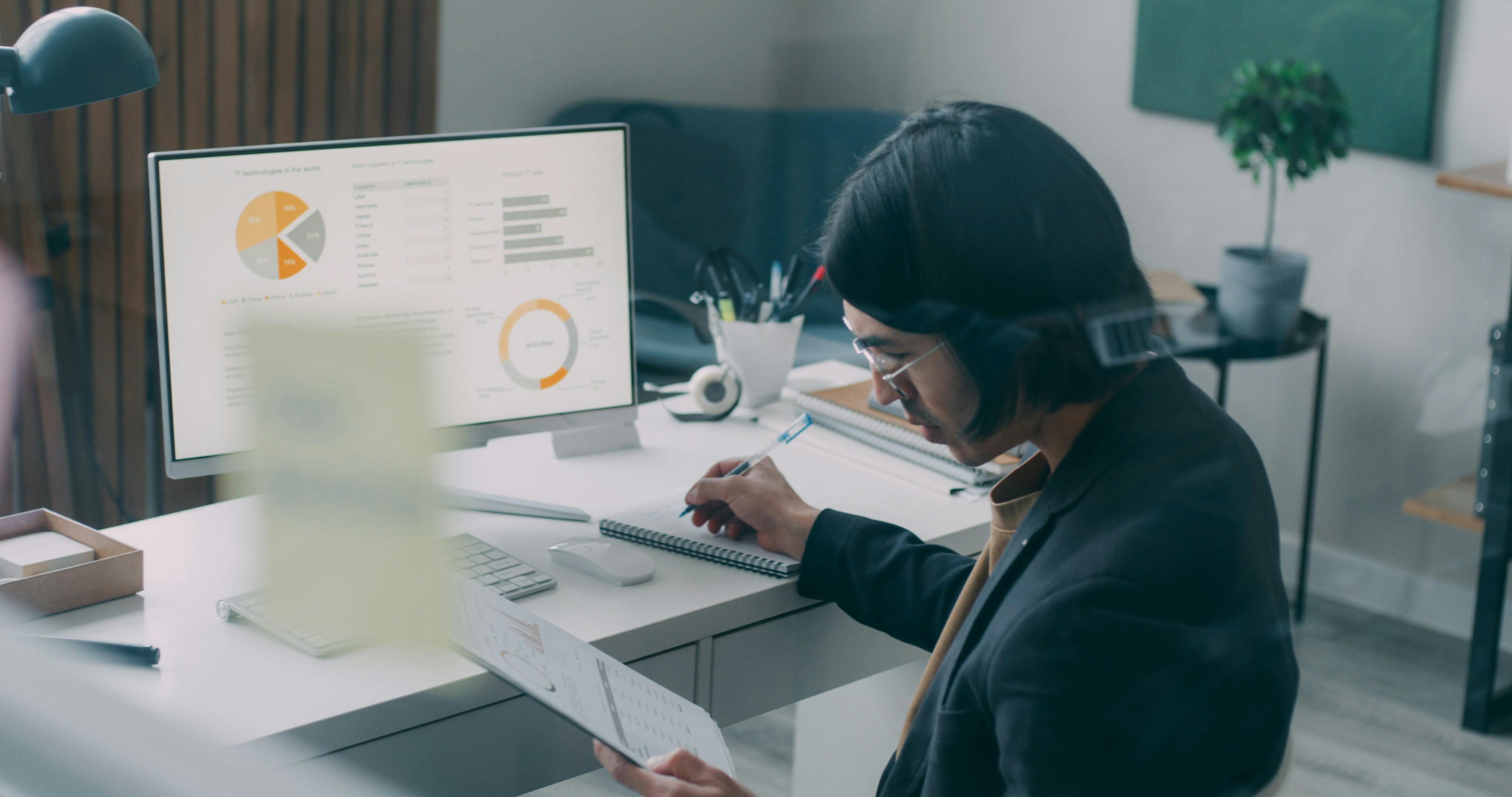 A woman wearing glasses and a dark blazer sits at a home office desk, writing in a notebook while holding a document and wearing a headset. A monitor behind her displays financial dashboard charts including pie charts and bar graphs in orange and grey.