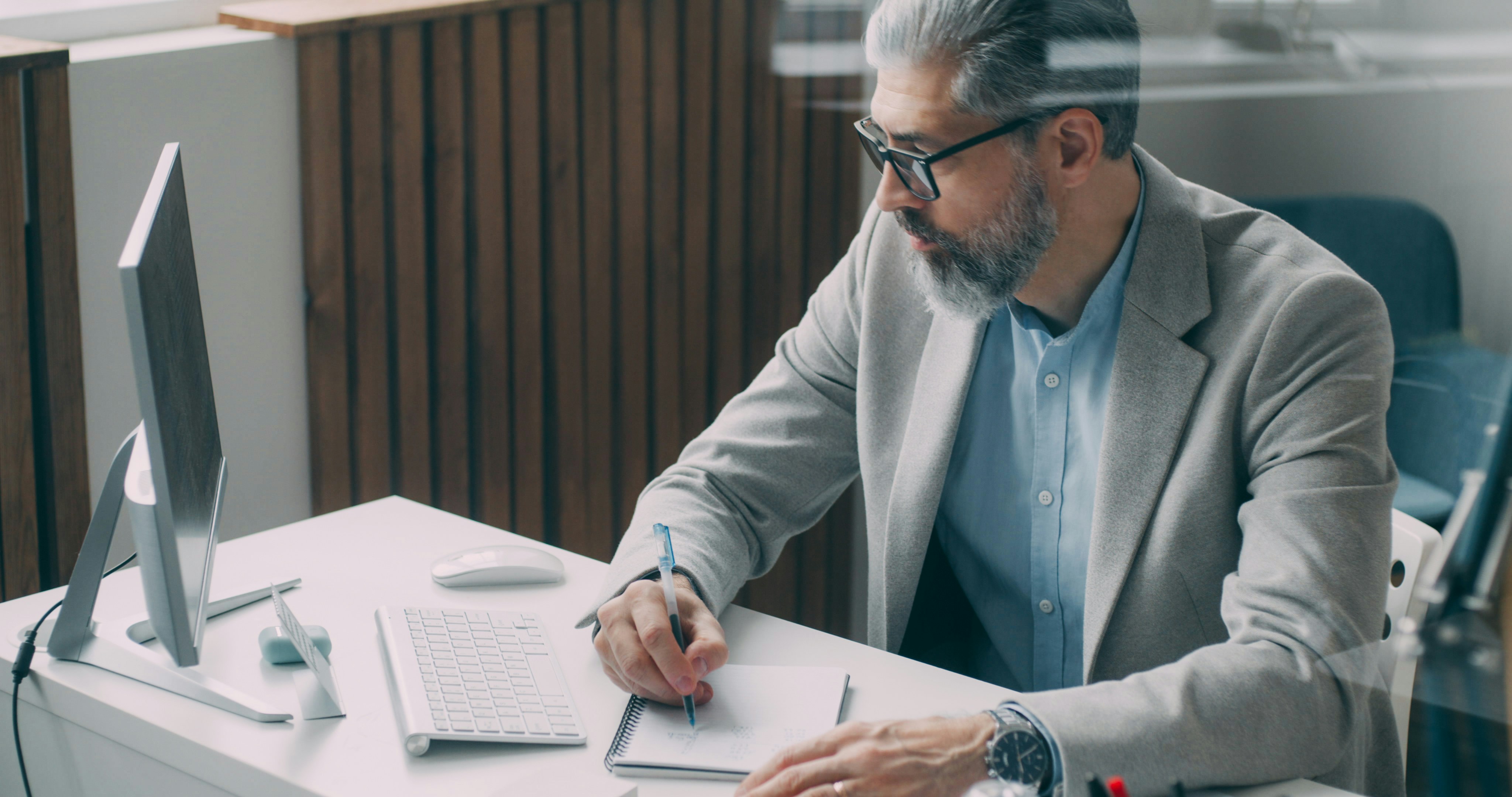 A middle-aged man with grey hair, a beard, and glasses wears a light grey blazer and blue shirt while writing in a notebook at a desk with a desktop computer, keyboard, and mouse visible in a modern office setting.