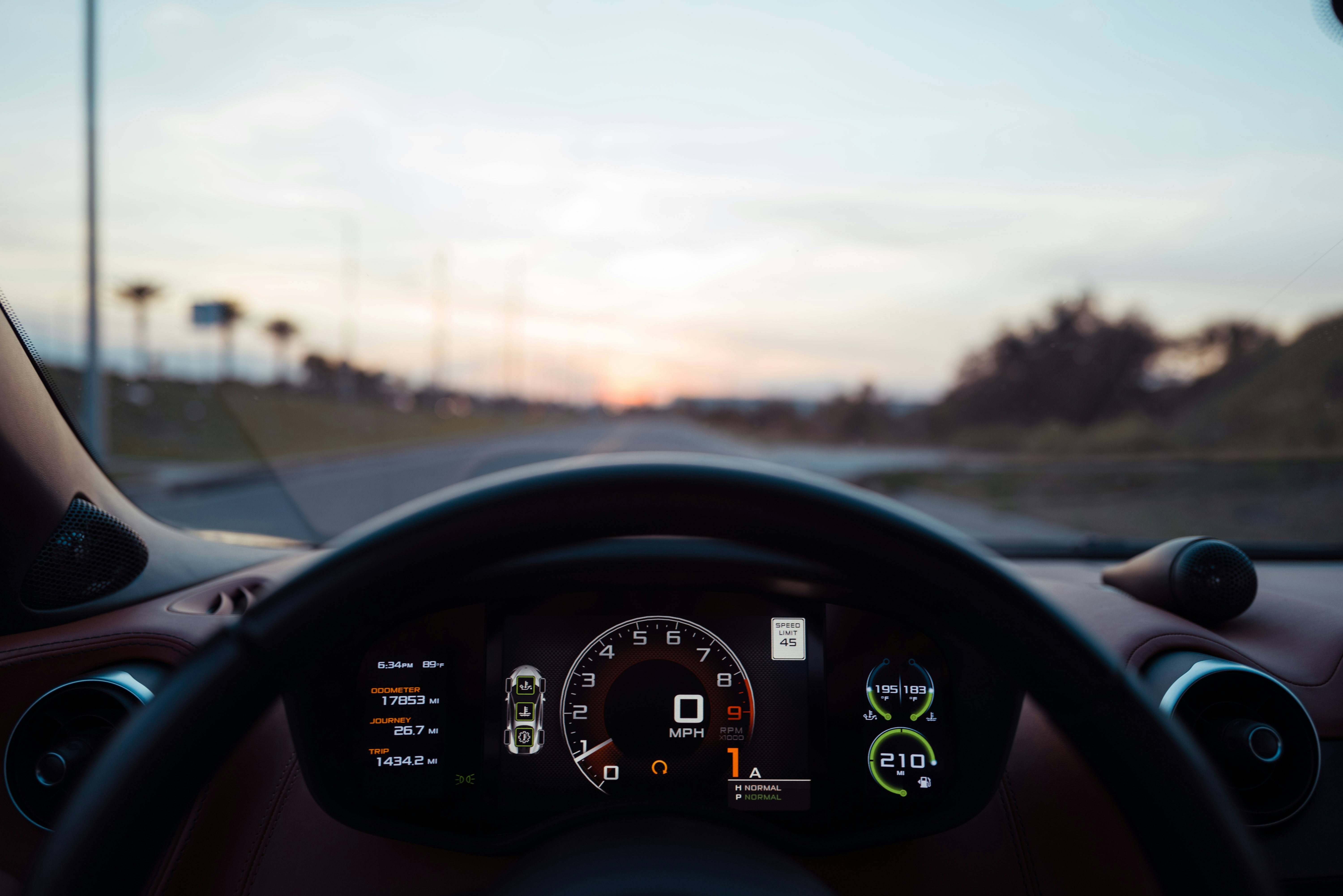 A view from inside a car's cockpit showing a digital instrument cluster with speedometer, RPM gauge, and trip information, looking out through the windshield toward an open highway at dusk with palm trees lining the road.