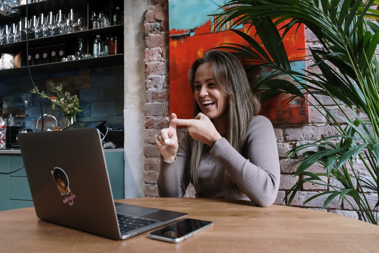 A woman with long brown hair laughs expressively during a video call on a laptop in a warm cafe setting with brick walls, colorful artwork, and a palm plant. The image conveys the positive and engaging experience of connecting with clients or employers through Somewhere's remote hiring platform.