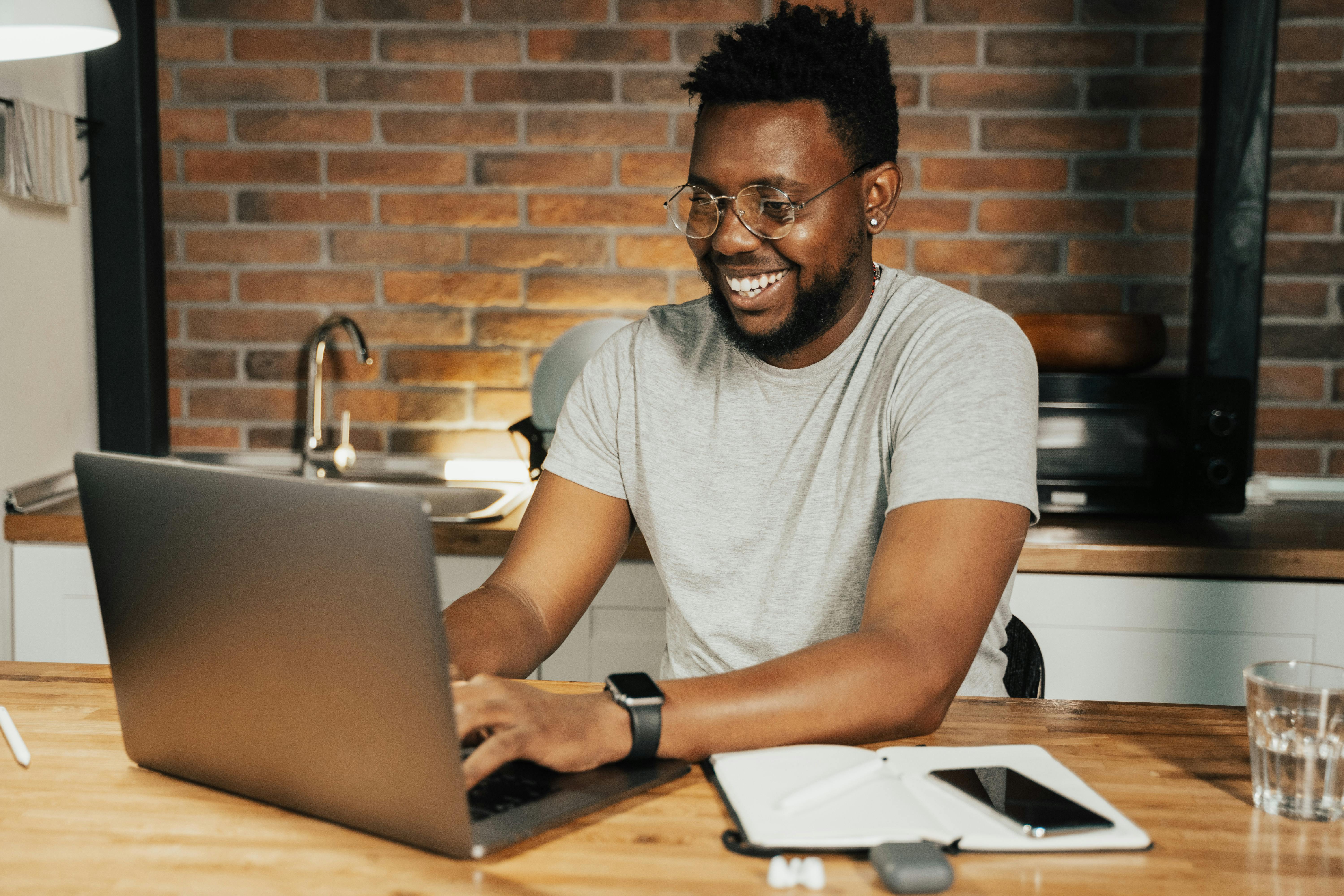 A smiling Black man with glasses and an earring works on a laptop at a wooden kitchen table in a home with brick walls. A smartwatch, tablet, and glass of water are visible nearby, depicting a happy and productive remote employee benefiting from flexible work arrangements.