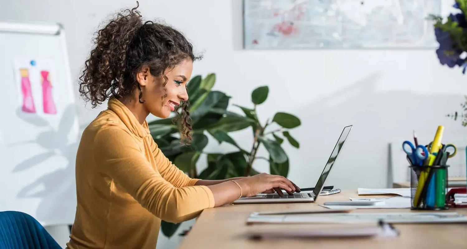 A smiling content writer with curly hair works on a laptop at a bright, modern office desk. A whiteboard with sketches is visible in the background alongside a large green plant, stationery holder, and framed art, conveying a creative and productive workspace.