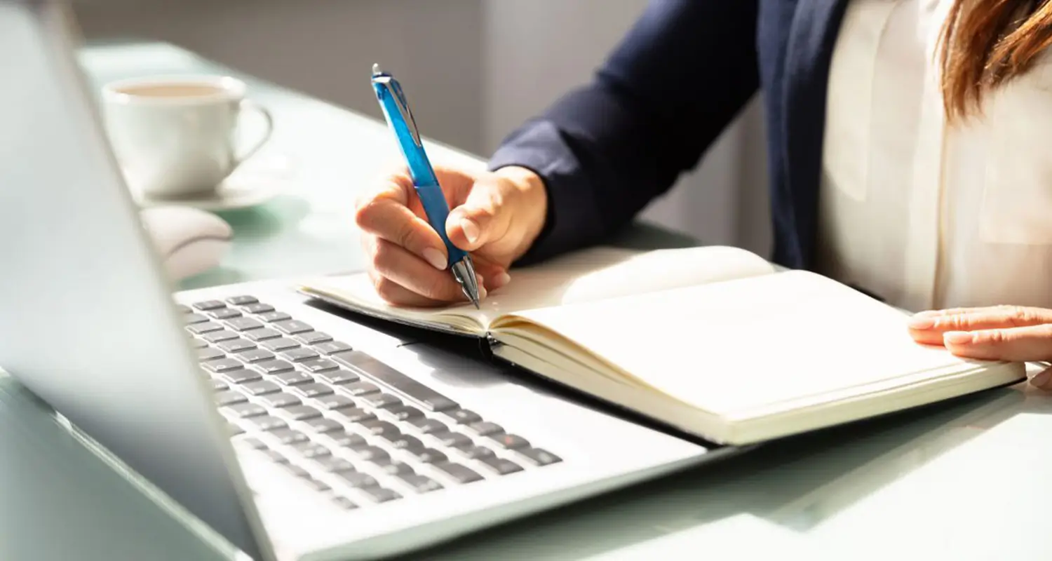 Close-up of a content writer's hands writing notes in an open notebook placed on top of a laptop keyboard, with a cup of coffee and a second open notebook visible nearby. The scene captures the research and planning phase of the content writing process.
