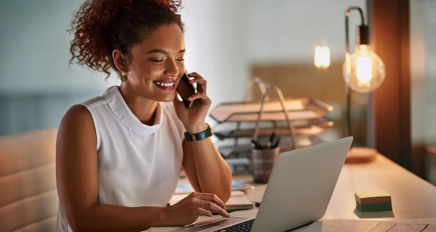 Smiling support professional talking on a phone while working on a laptop in a warmly lit office.