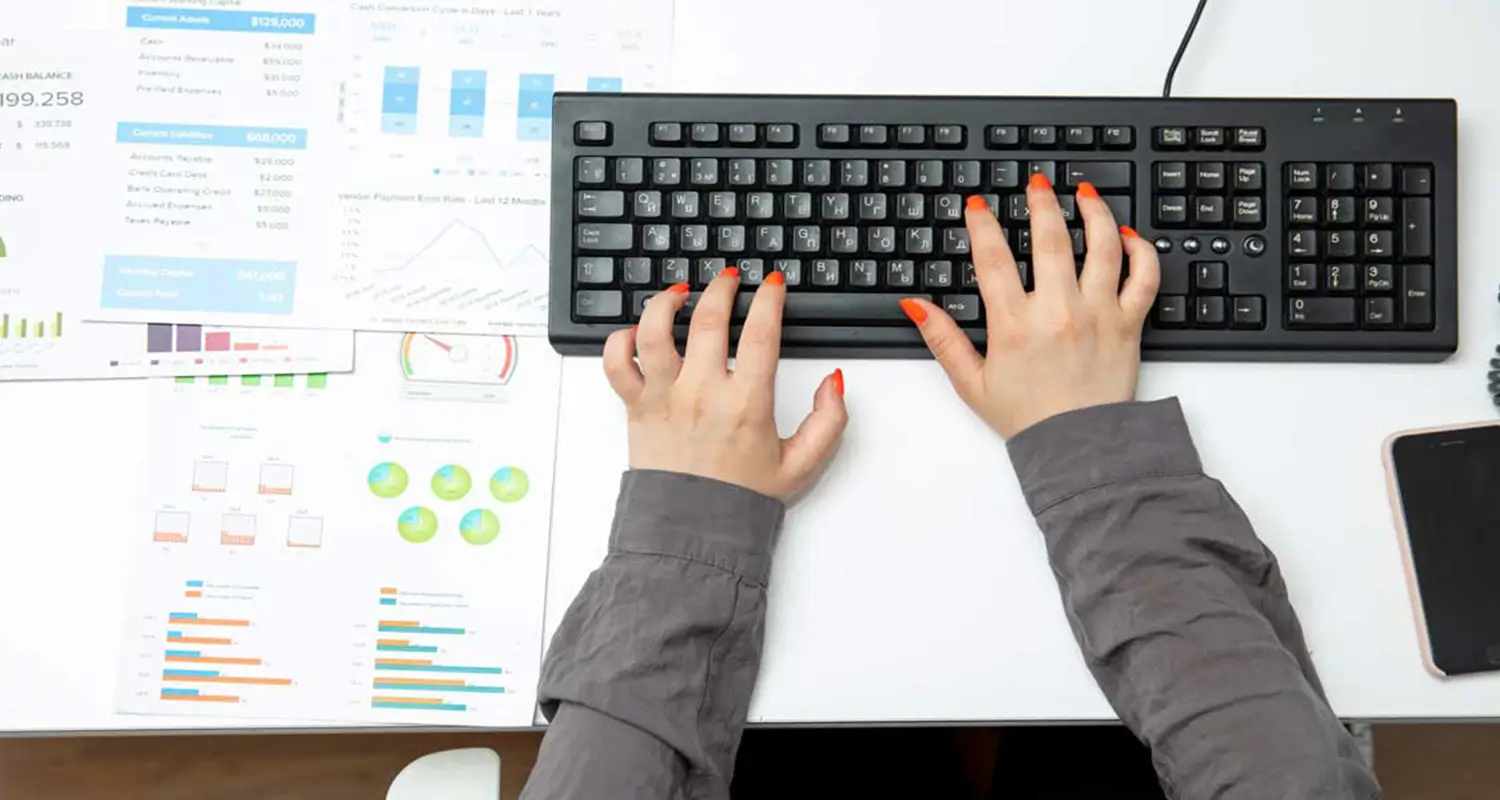 Close-up of hands typing on a keyboard surrounded by printed financial reports and data charts