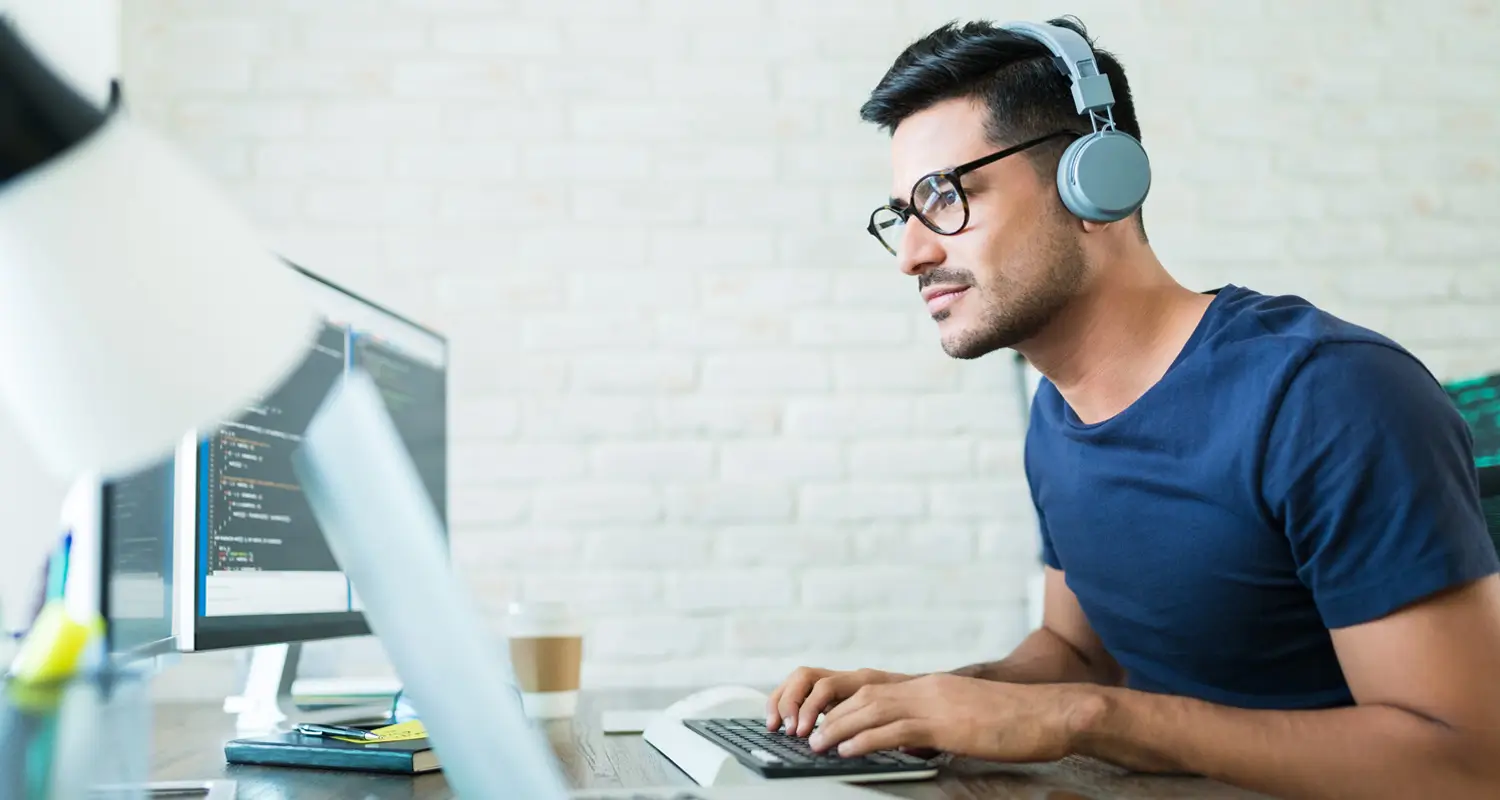 Male developer with headphones and glasses focused on dual monitors showing code at a modern white brick office desk.
