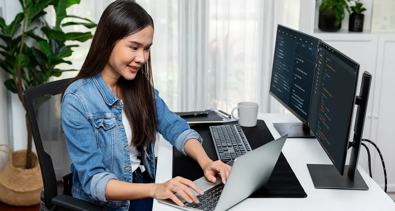 Female developer in a denim jacket working on a laptop with dual monitors displaying code in a bright home office.