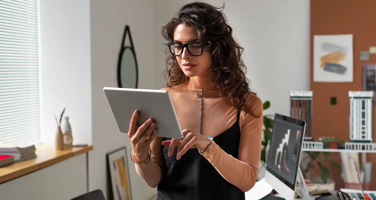 Marketing professional in glasses browsing a tablet in a creative studio with a desktop monitor in the background.