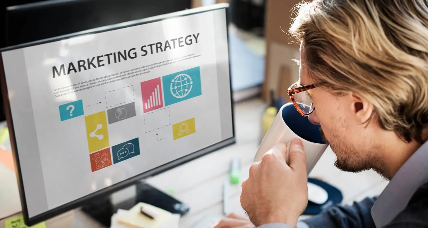 Professional reviewing a marketing strategy presentation on a large monitor while drinking coffee at a desk.