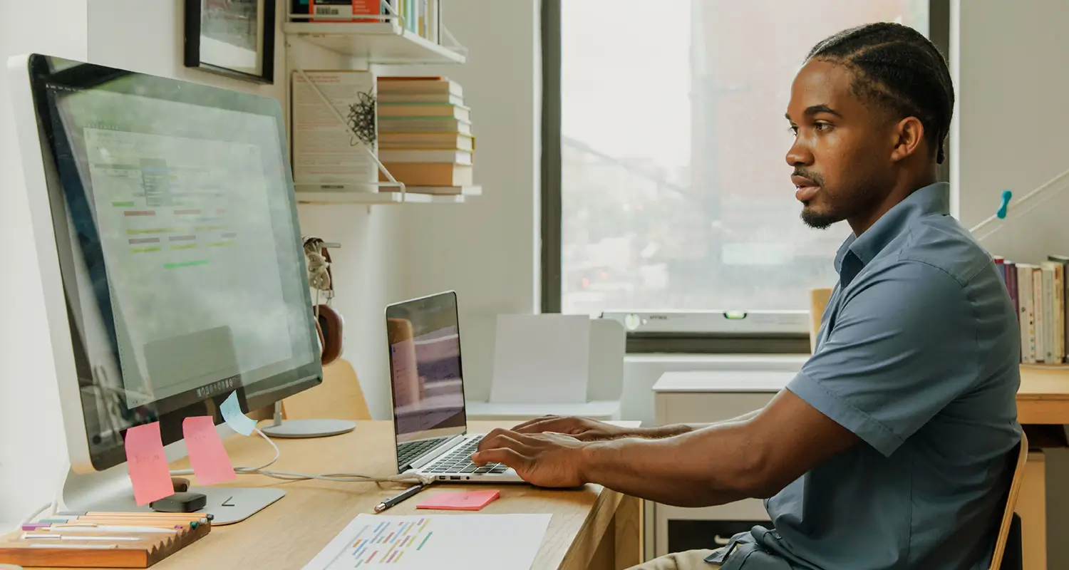 A male designer works at a home office desk with a large monitor displaying colorful timeline or spreadsheet data and an open laptop beside it. Bookshelves, pink sticky notes, and a casual workspace setting reflect a focused remote work environment.