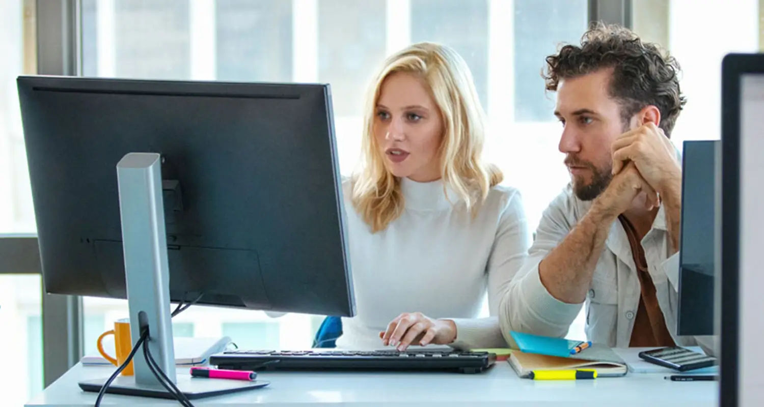 A female marketing designer with blonde hair presents campaign visuals to a male colleague as both focus on a desktop monitor in a bright, modern office. Markers and sticky notes are scattered on the desk, suggesting an active creative collaboration session.