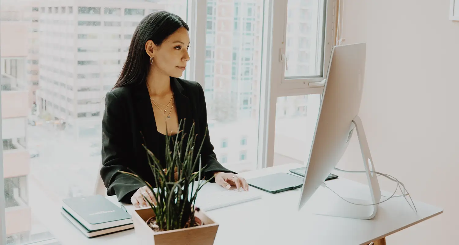 Paralegal in a black blazer works at a desktop computer in a bright office with a city view
