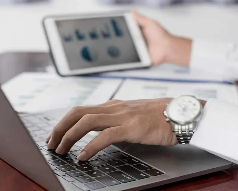 Professional reviewing analytics charts on a tablet while typing on a laptop, with printed reports spread on the desk.