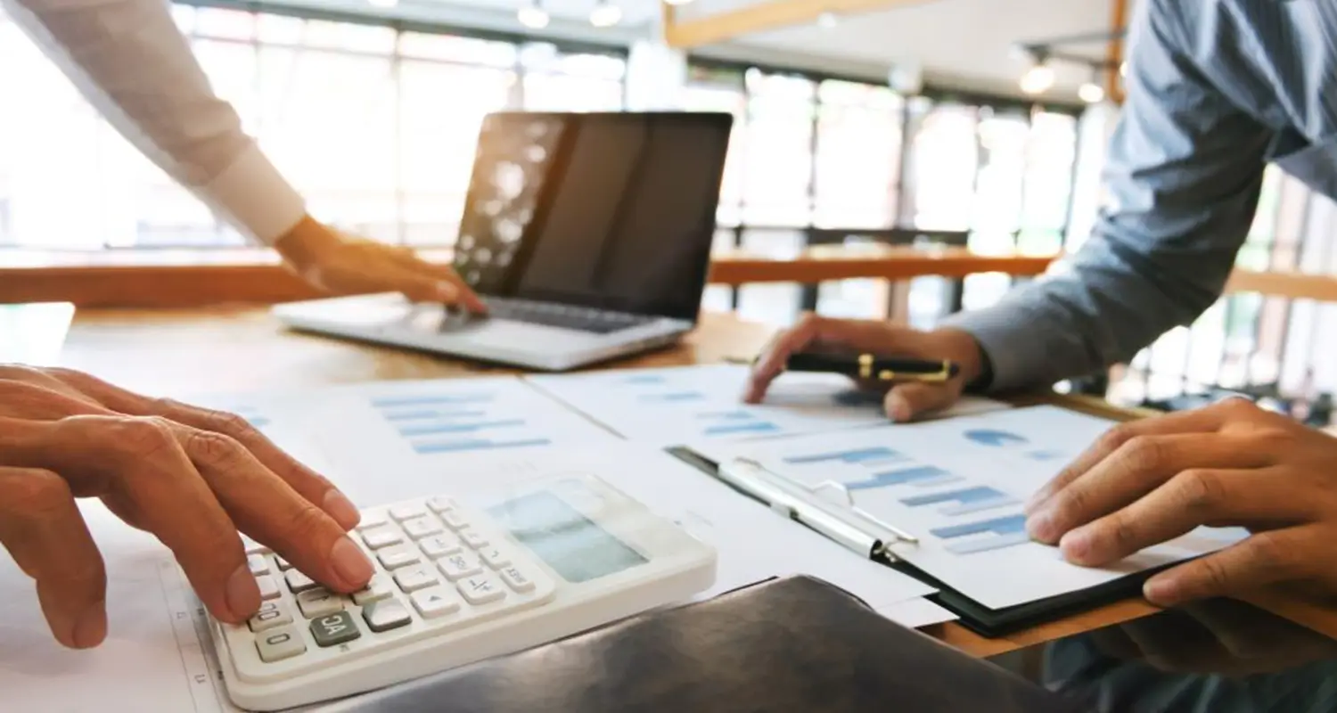 Two business professionals collaborate at a desk covered with printed charts, bar graphs, and financial reports. One person operates a calculator while the other holds a pen and reviews a clipboard with data, depicting a quote coordinator and colleague working through pricing analysis and margin calculations.