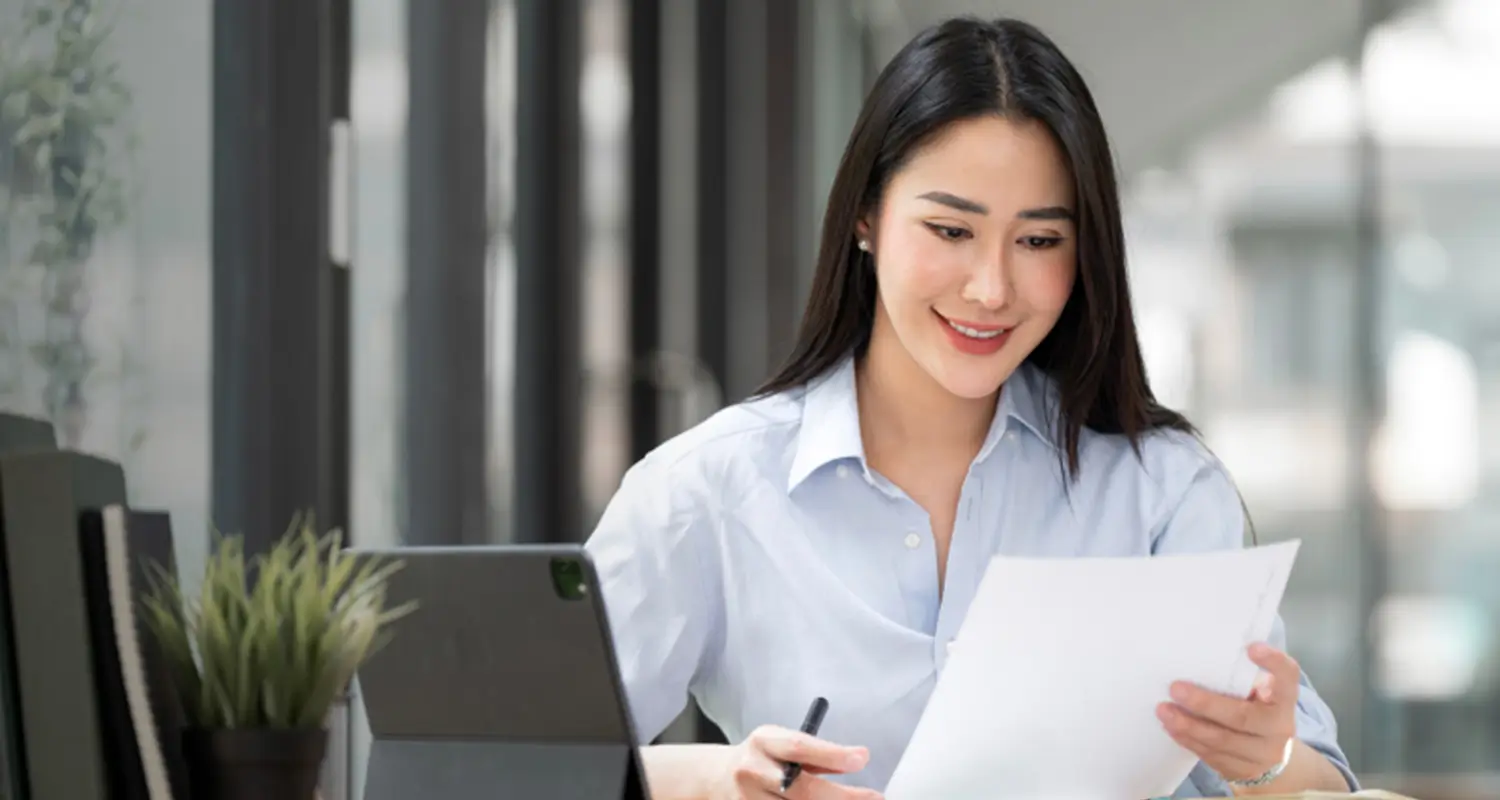 A smiling woman in a light blue button-up shirt reviews printed documents while sitting at a desk with a tablet beside her in a modern, glass-walled office. She holds a pen and appears to be carefully checking quotes or proposals, representing a quote coordinator managing deal documentation.