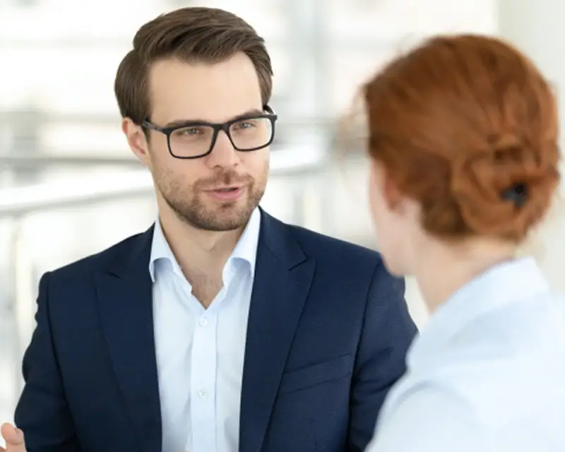 Man with glasses in a navy suit speaking with a red-haired woman in a professional office setting