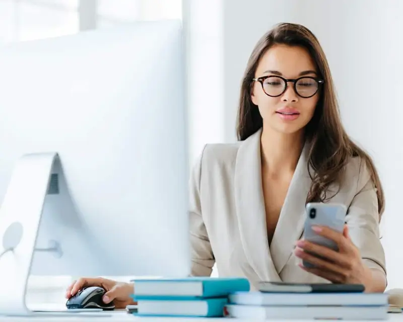 Woman with glasses at a desk checking her phone while using a computer mouse, with books nearby