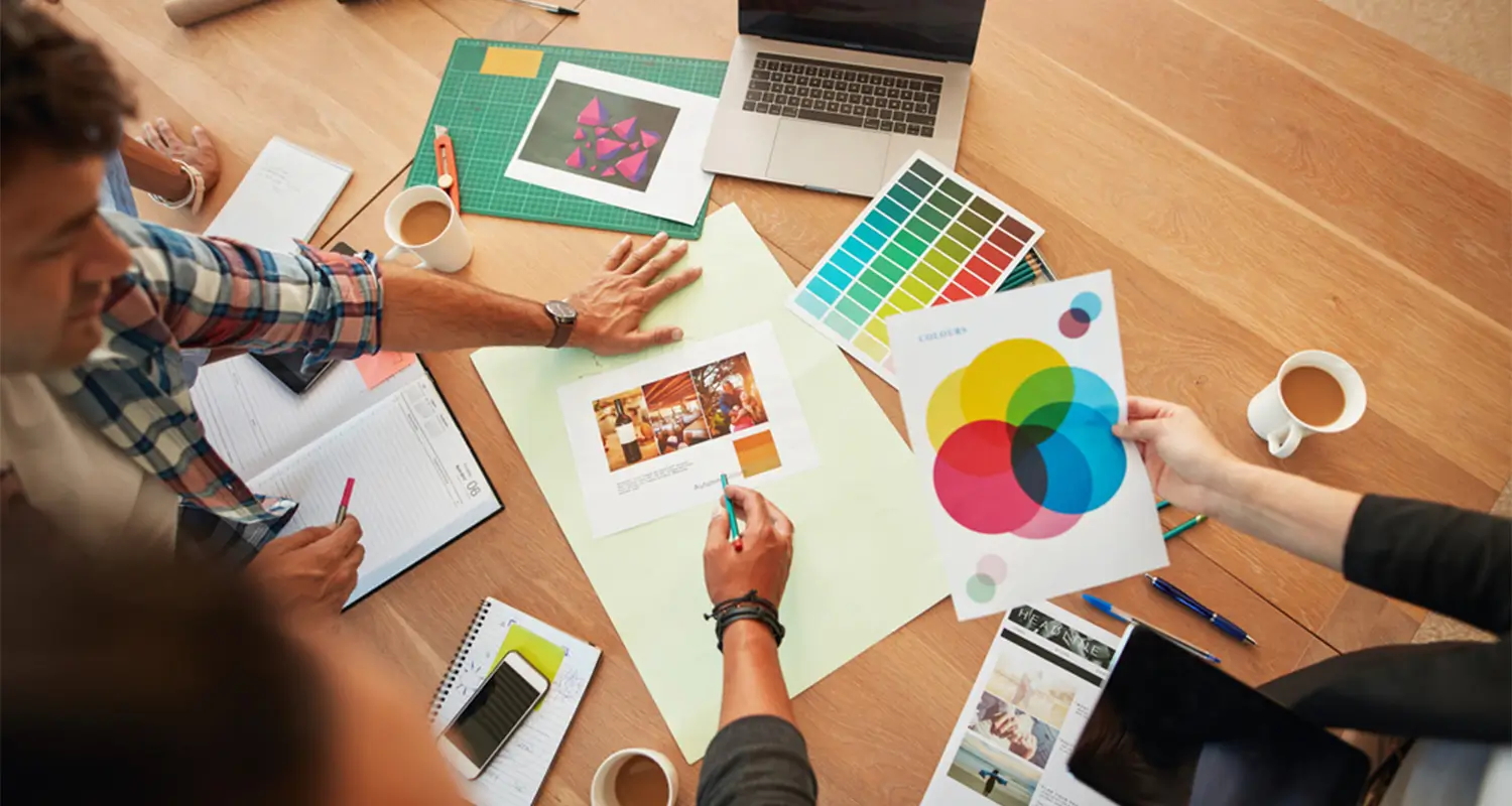 Overhead view of a creative team brainstorming around a wooden table covered in color theory charts, Pantone swatch fans, printed mood board photos, notebooks, and coffee cups. A laptop sits open at the top and one team member holds a color wheel diagram showing CMYK overlapping circles.