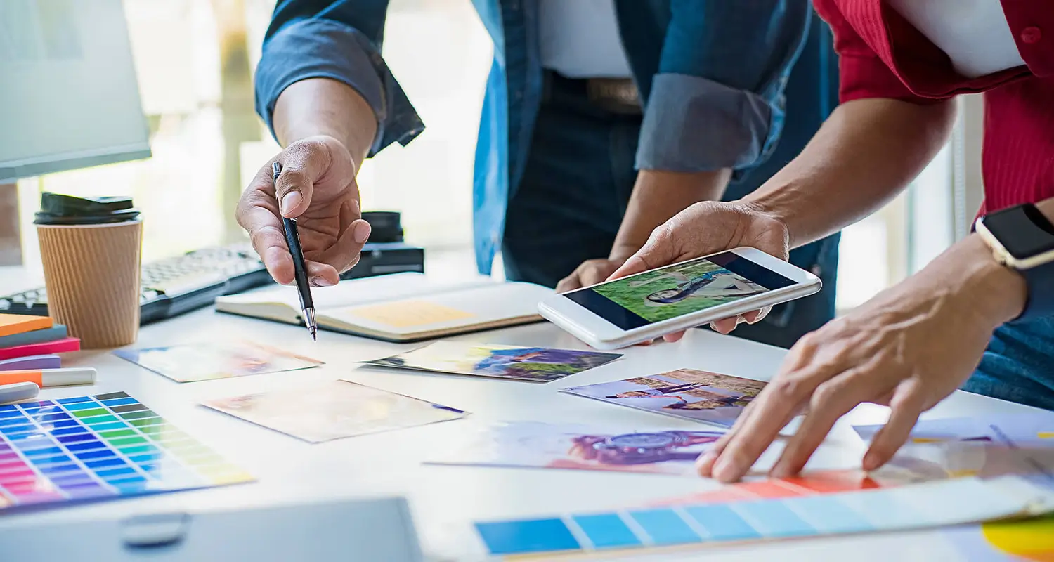 Two advertising designers review campaign materials at a desk covered in Pantone color swatches, printed photo references, and a notebook. One holds a smartphone showing a lifestyle photo while the other points with a pen, collaborating on visual direction for an ad campaign.
