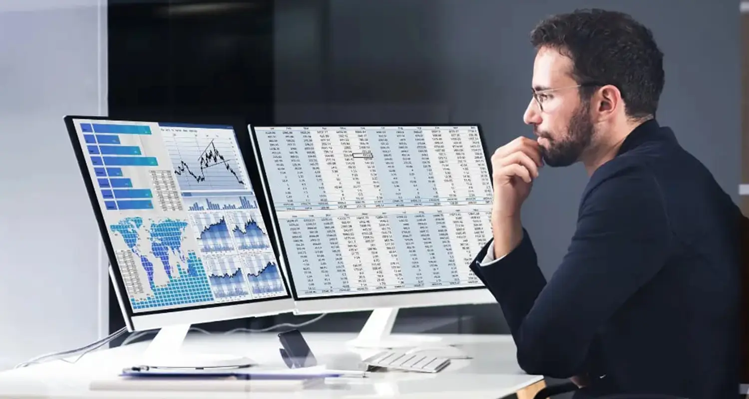 Man with glasses studying dual monitors displaying analytics dashboards with world map heatmaps, line charts, and spreadsheet data