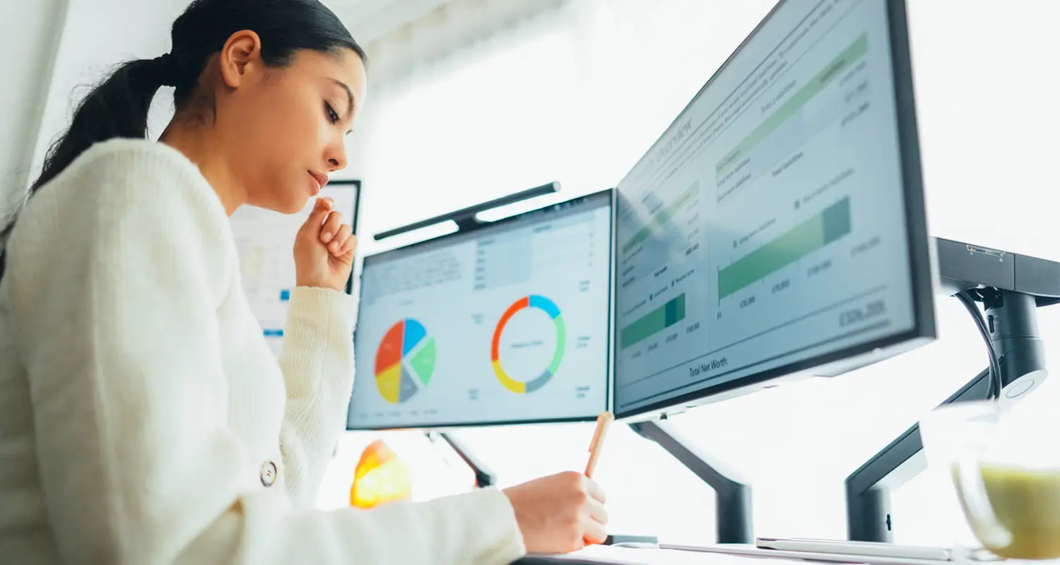 Woman in a white sweater thoughtfully reviewing dual monitors showing colorful pie charts and financial dashboards while taking notes