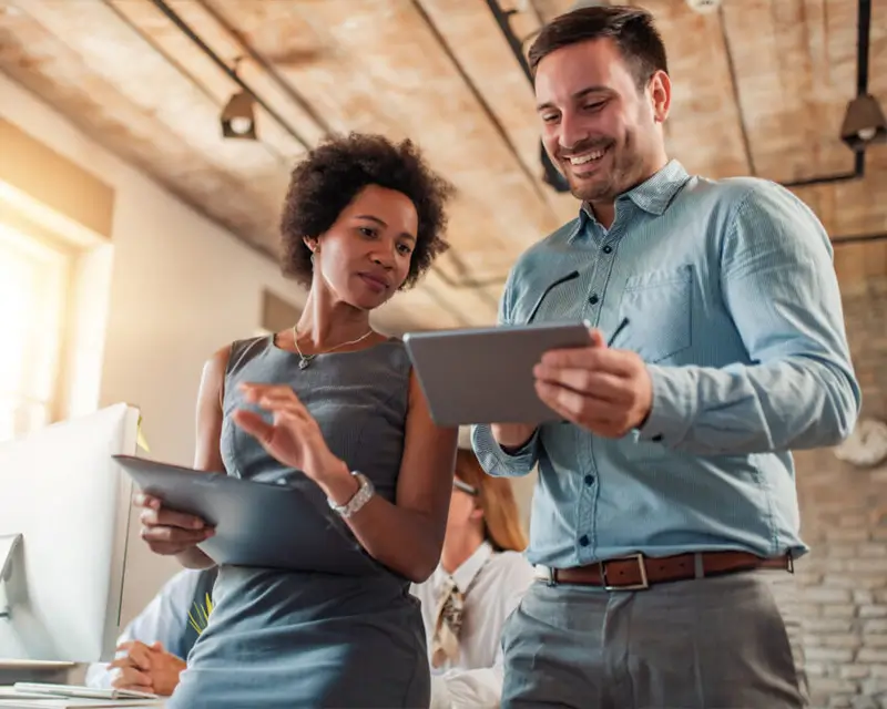 Two colleagues smiling and reviewing content on a tablet together in a modern open office space