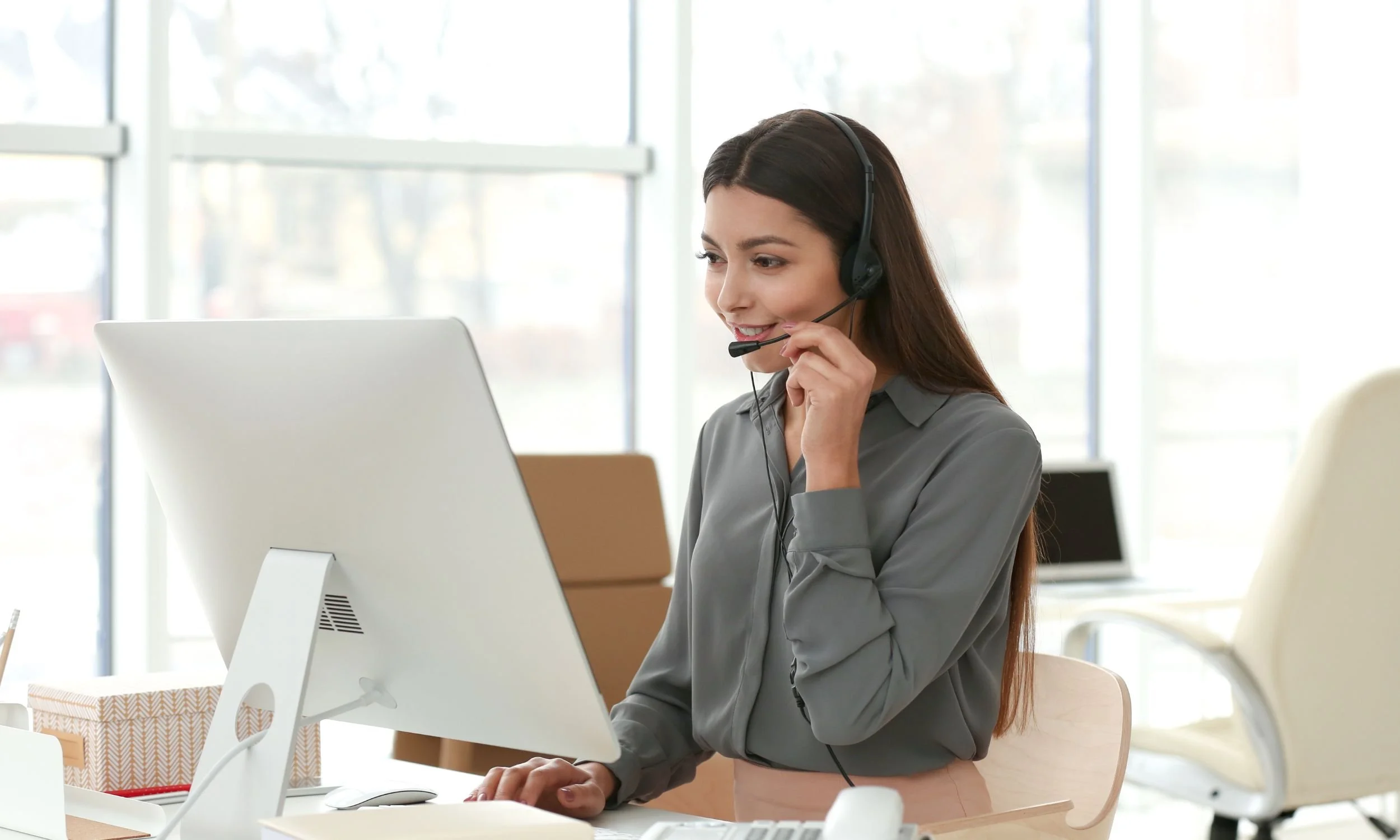 Woman wearing a headset smiling while working at a desktop computer in a bright, modern office