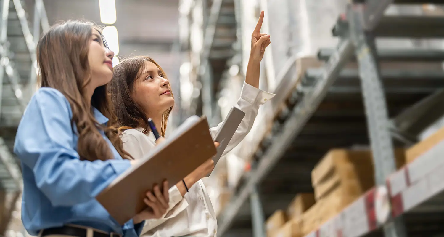 Two warehouse employees review inventory on shelves using a clipboard, pointing at stock levels