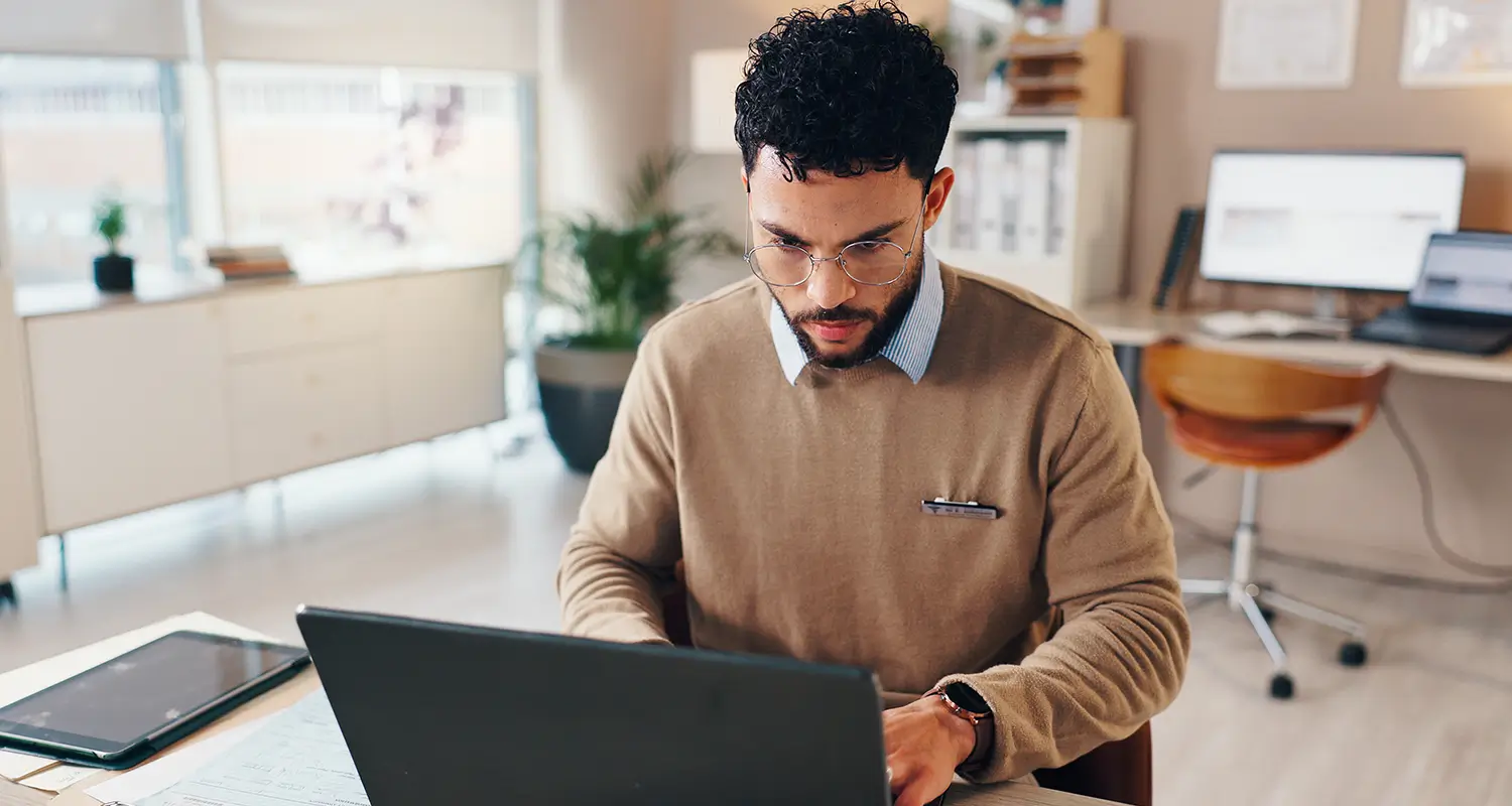 Man in glasses and sweater focused on typing on a laptop in a bright modern office