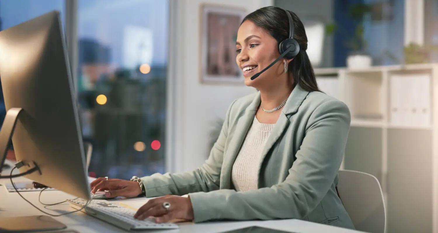 Smiling operations professional wearing a headset while working at a desktop computer in an office at dusk.