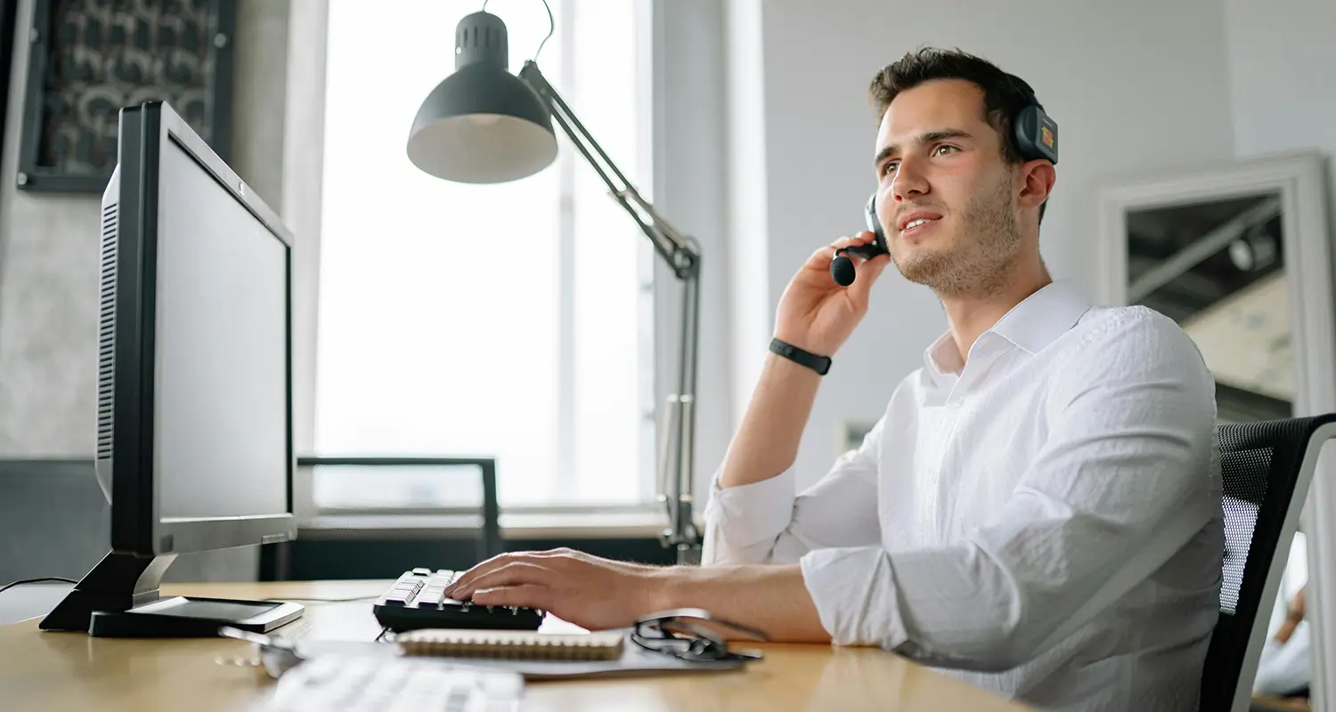 Male operations professional wearing a headset at a desk with a monitor and desk lamp in a bright modern office.