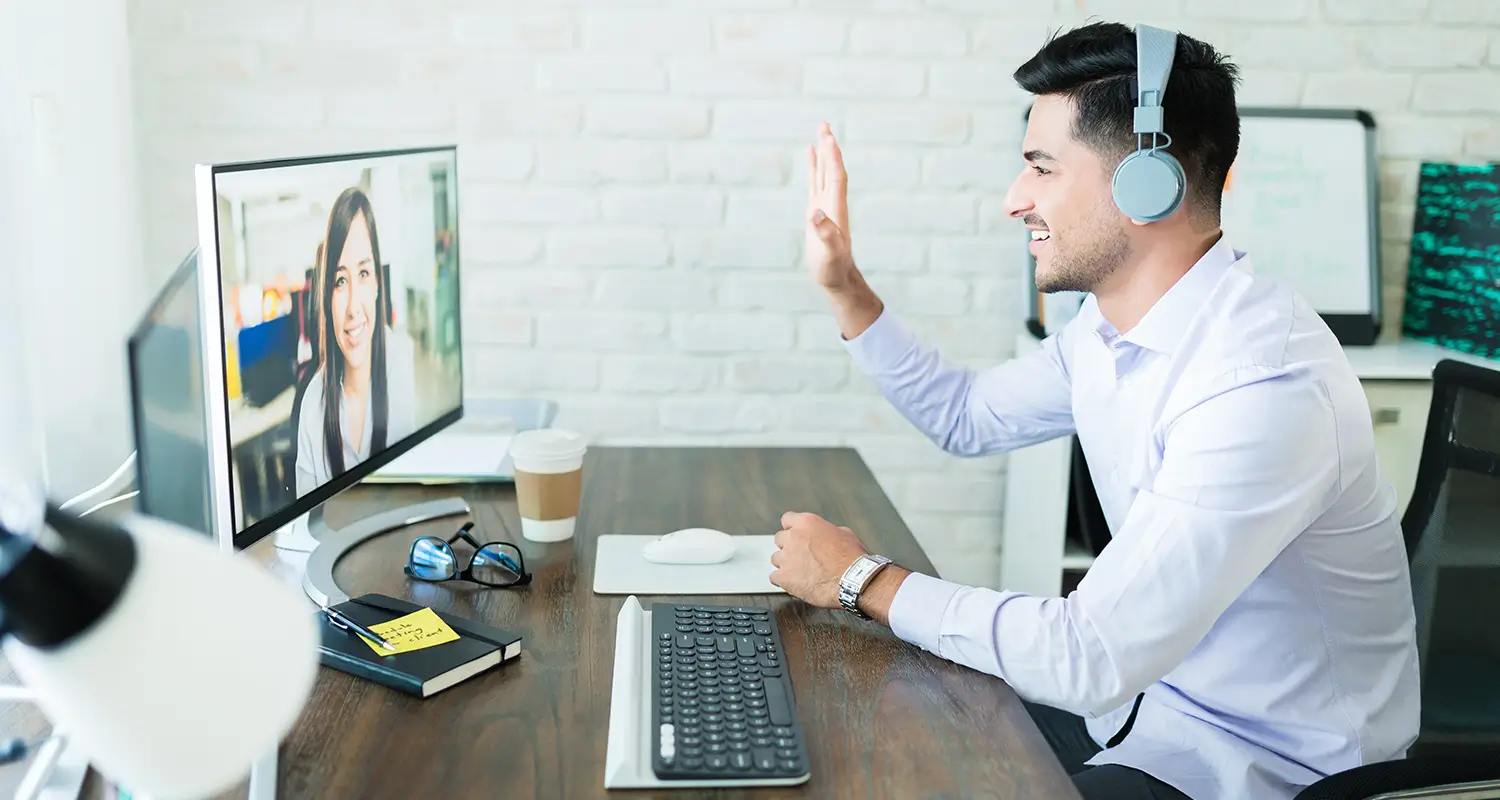 Man with headphones waving at a remote colleague on a desktop monitor during a video call, with sticky notes and glasses on the desk.