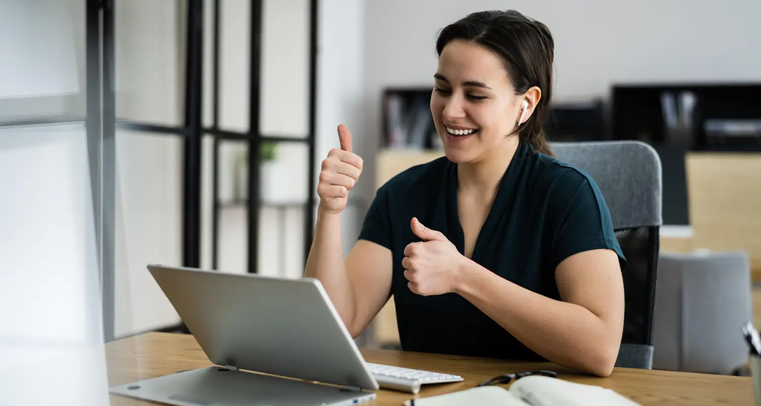 Smiling woman giving two thumbs up during a video call on a laptop, wearing an earpiece, seated at a wooden desk in a bright office.