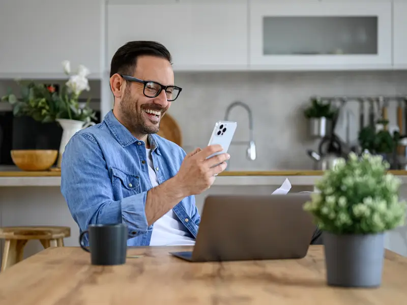 A smiling man with glasses and a denim shirt laughs while looking at his smartphone, seated at a kitchen table with a laptop open in front of him. A small plant and a smart speaker are on the table, depicting a relaxed and connected remote work lifestyle.