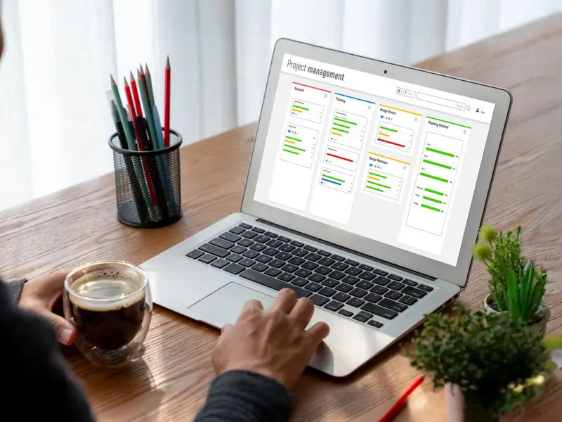 A person holds a glass of espresso while browsing a project management dashboard on a laptop screen. The interface displays columns labeled Research, Planning, Design Review, and Planning Finished, with color-coded task bars, illustrating how remote-first teams organize workflows digitally.