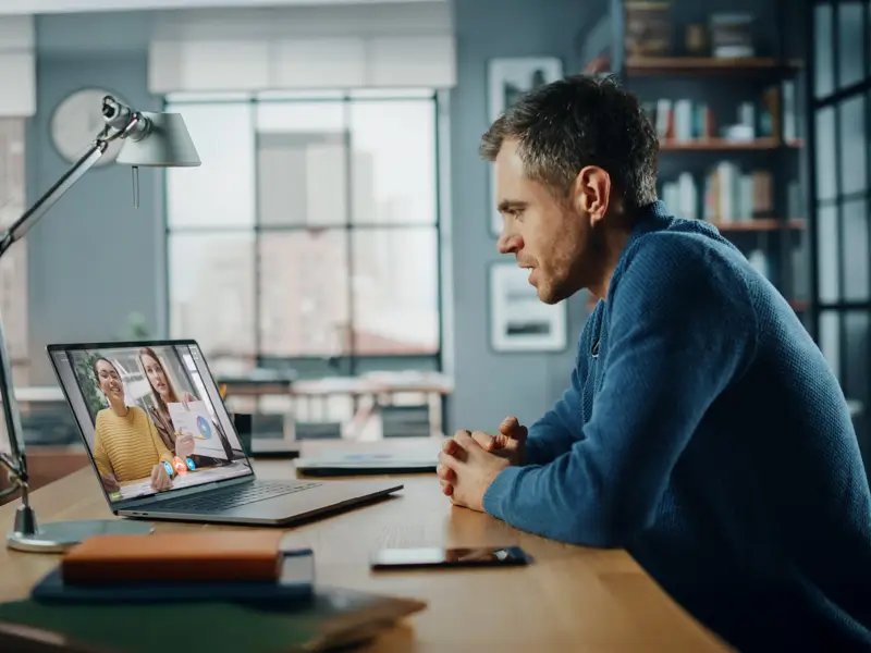 A man in a blue sweater sits at a wooden desk in a modern loft-style home office, leaning forward with hands clasped while watching a video call on his laptop. The screen shows two women, one holding a chart, representing a remote team collaboration session.