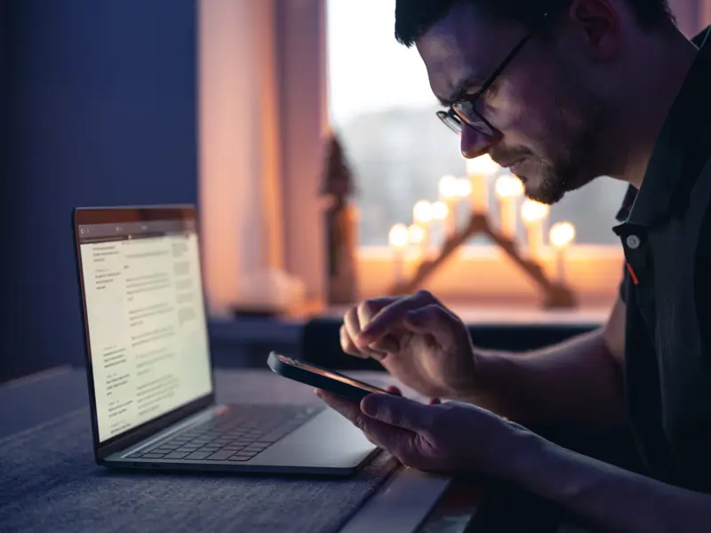 Man with glasses using a smartphone beside a laptop in a dimly lit room with candles in the background