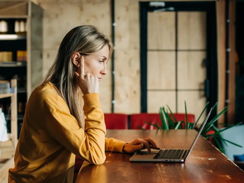 Woman in a yellow sweater focused on her laptop at a long wooden table in a modern workspace