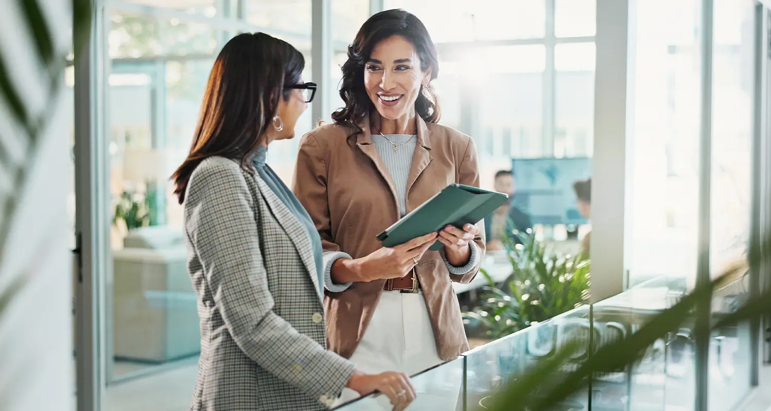 Two professional women in an office hallway smiling and discussing while one holds a tablet, with plants in the background