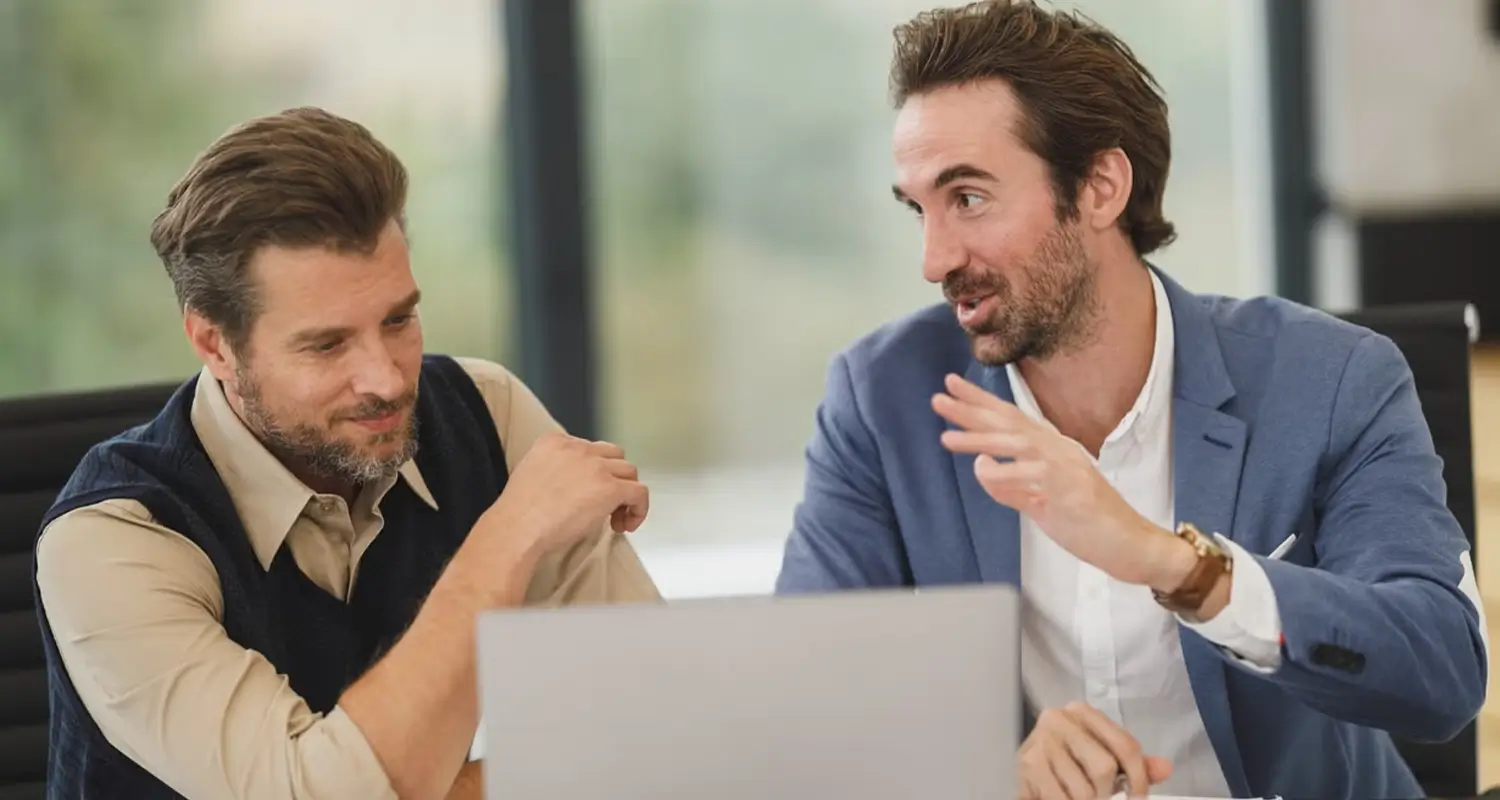Two men in business casual attire discussing something at a laptop in a bright modern office, one gesturing expressively