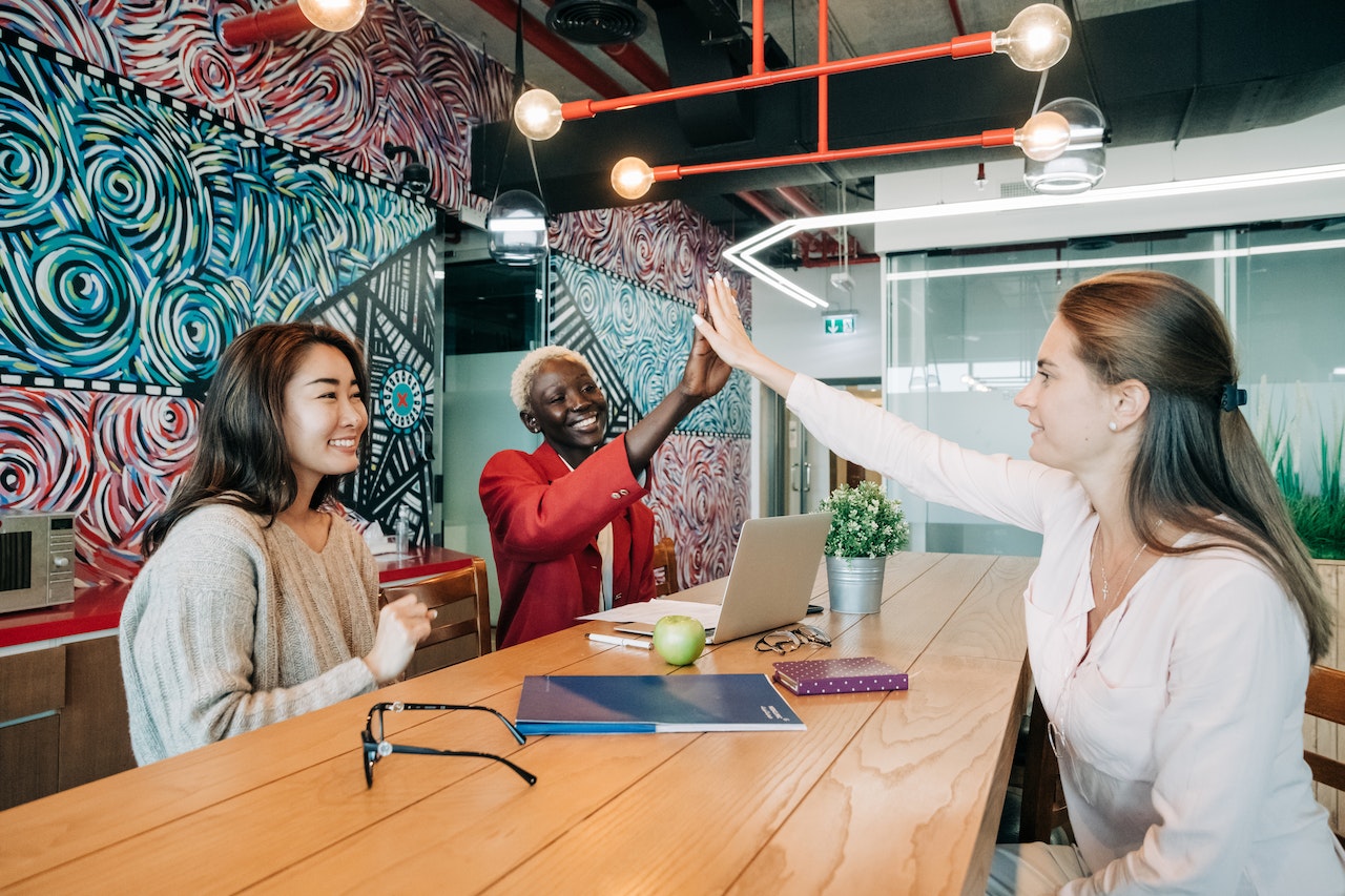 Three women in a colorful creative office space celebrating together. One woman in a red blazer gives a high five to a colleague in white while a third woman in a knit sweater smiles nearby. A laptop, notebooks, and a small plant sit on the table, conveying a positive team dynamic.