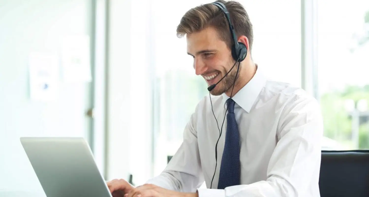 A smiling man in a white dress shirt and navy tie wears a headset while working on a laptop in a bright, modern office. He represents a professional remote customer service representative handling customer inquiries with a positive demeanor.