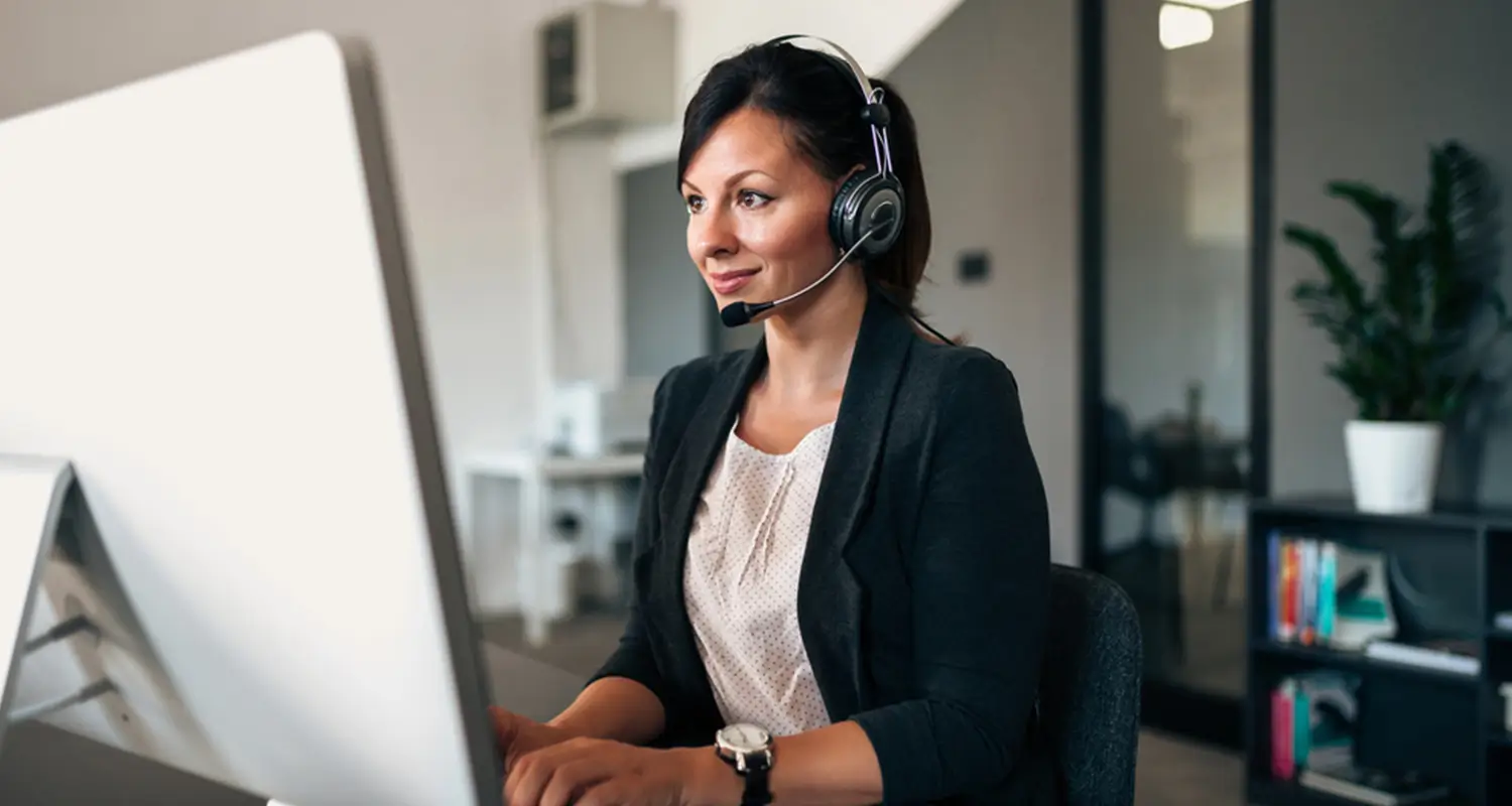 A woman in a black blazer and headset looks attentively at a desktop monitor while working in a sleek open-plan office. A bookshelf and potted plant are visible in the background, depicting a focused remote customer service agent managing support interactions.