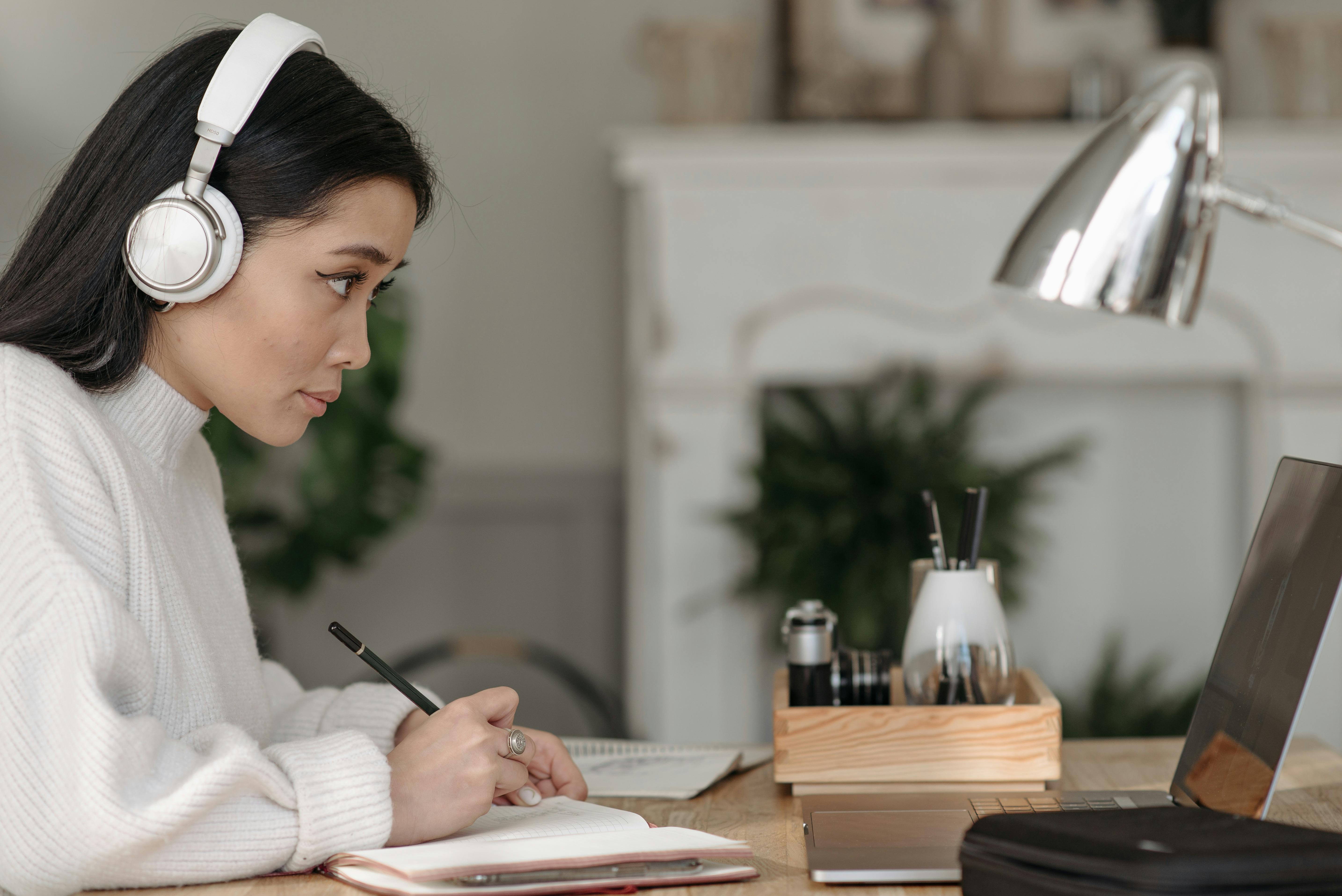 A woman wearing white over-ear headphones writes in a notebook at a well-organized home desk with a lamp and laptop. Her focused expression and note-taking posture suggest she is participating in a remote interview or online training session.