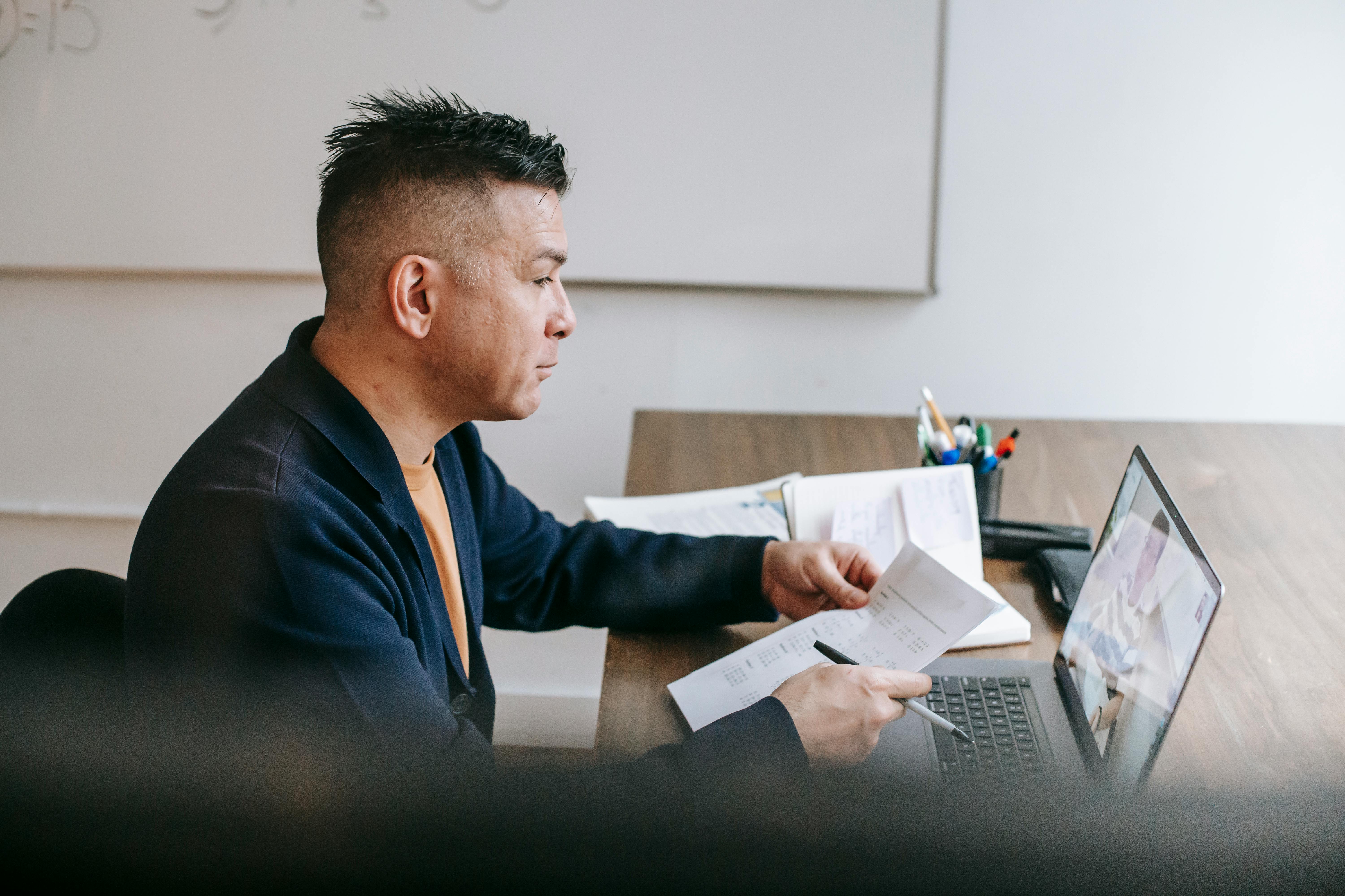 A man in a dark blazer sits at a conference table reviewing handwritten notes while participating in a video call on a laptop. A whiteboard and open notebooks are visible in the background, suggesting a remote hiring or interview session.